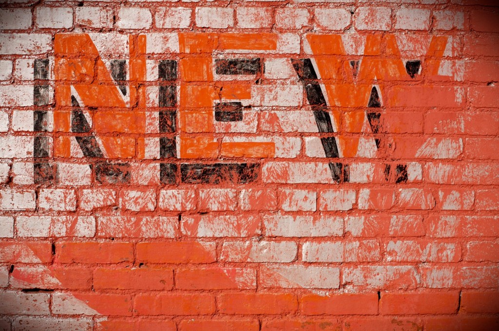 The word NEW painted in orange with black shadow on a brick wall. The brick wall is painted in orange with an overlay of white.